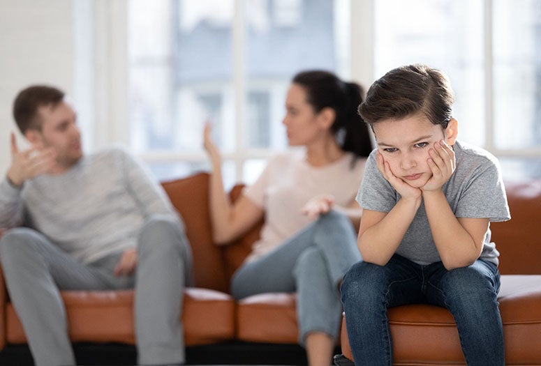 Child sitting sadly on sofa with parents arguing in the background. 