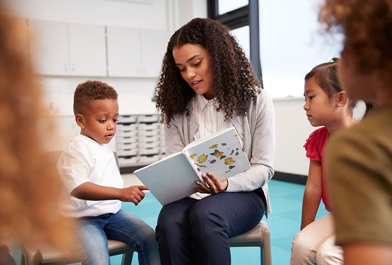 Infant school boy pointing in a book held by the female teacher, sitting with kids in a circle on chairs in the classroom, close up
