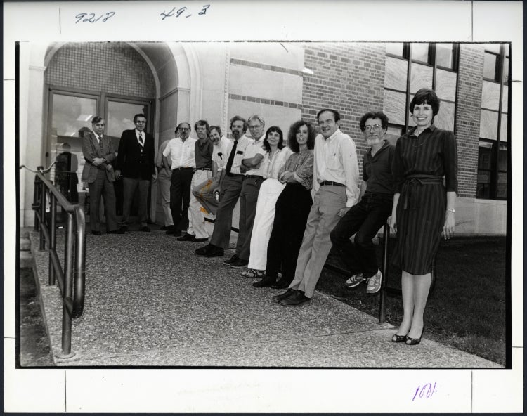 The faculty of Rice University’s Department of English, in a photograph used in the 1987 Campanile Yearbook. They are shown at one of the ramped entrances to Rayzor Hall, with faculty standing in front of the arched doorway or leaning or sitting on the guard rail along the ramp. From left to right: William Piper, Alan Grob, Terrence Doody, John Meixner, Edward Snow, Edward Doughtie, Wesley Morris, Walter Isle, Meredith Skura, Susan Wood, Jack Ward, Max Apple, and Mary Tobin.