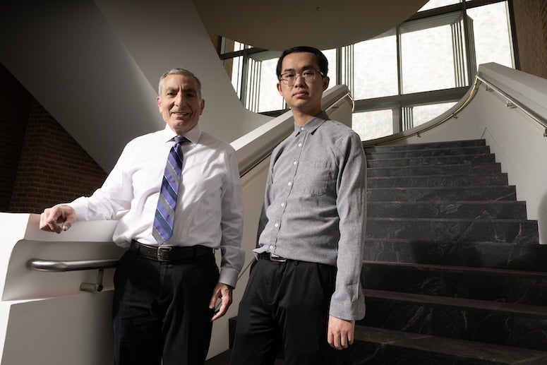 Two men, one older and white, one younger and Asian, stand at the bottom of a staircase