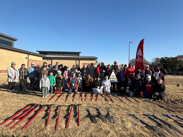 A group of people standing outside on a sunny day. A row of shovels and tools is laid out on the ground in front of them.
