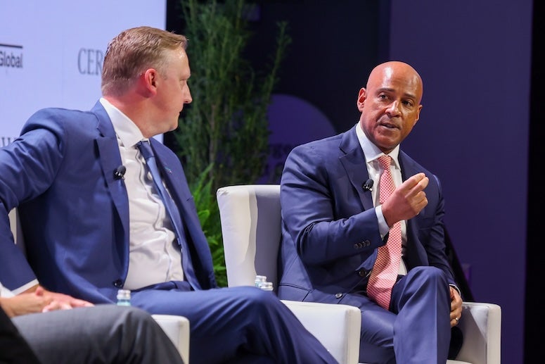 A white man, wearing a suit and sitting on a chair, listens as a Black man, wearing a suit and sitting on the chair next to him, talks.