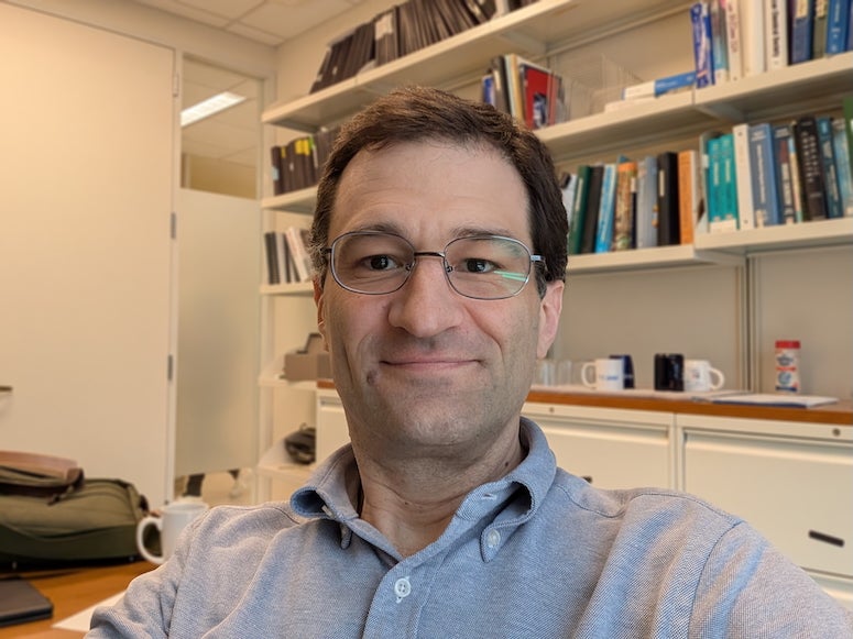 A white man smiles at the camera. He is sitting in front of a wall of books and wearing glasses.