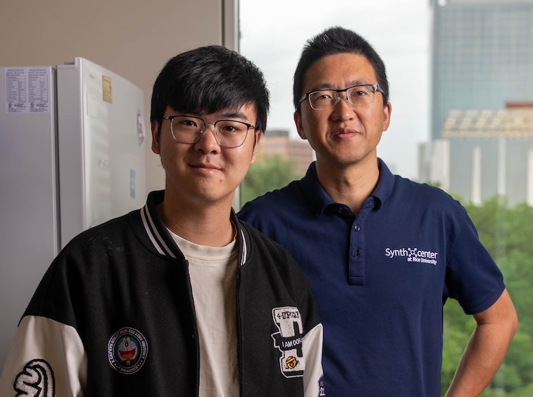 A young Asian man in a letterman jacket stands in front of a middle-aged Asian man in a polo shirt. Both are wearing glasses.