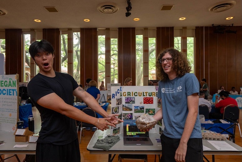 An Asian man excitedly points at a mushroom growth brick held by a laughing white man with curly hair