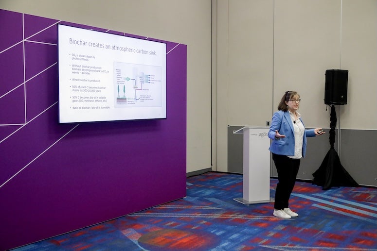 A white woman stands in front of a podium, presenting a slide deck projected on the wall behind her