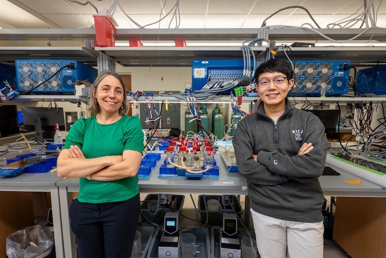 An older white woman and a young Asian man stand in front a lab bench with bottles and wires on it. They are smiling at the camera