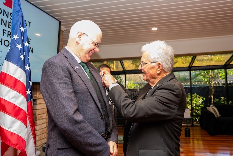 A tall white man stands and smiles at a shorter white man pinning something on his suit coat.