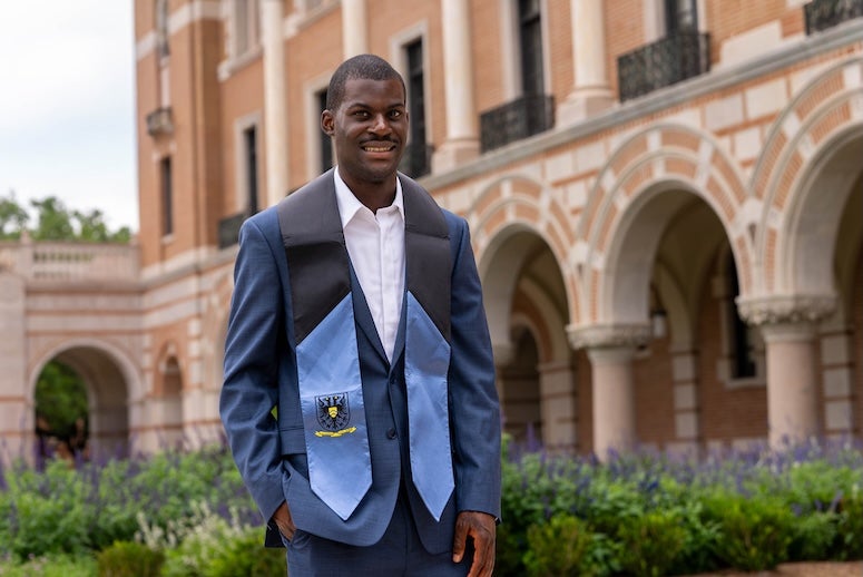 A Black man in a suit and blue stole stands in a university courtyard.