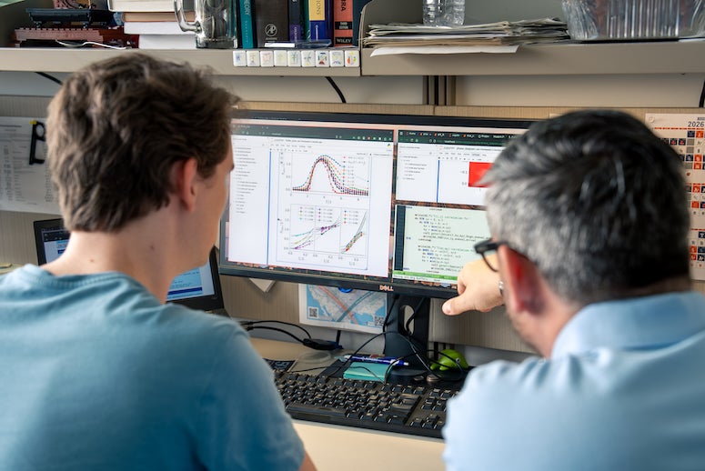 Two white men look at a computer screen which has graphs and datasets on it.