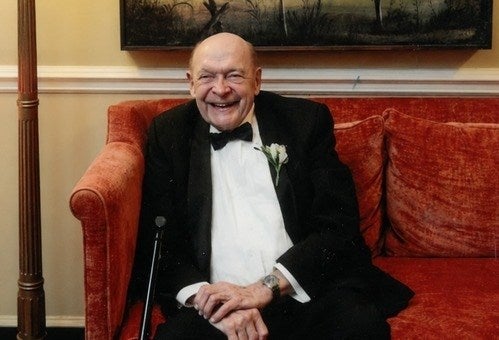 A grinning older white man sits on a red couch. 