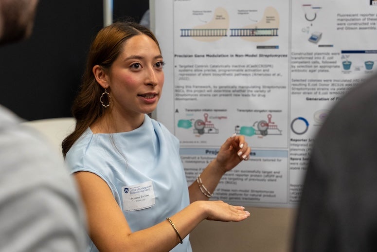 A young woman gestures at a poster while talking to an audience.
