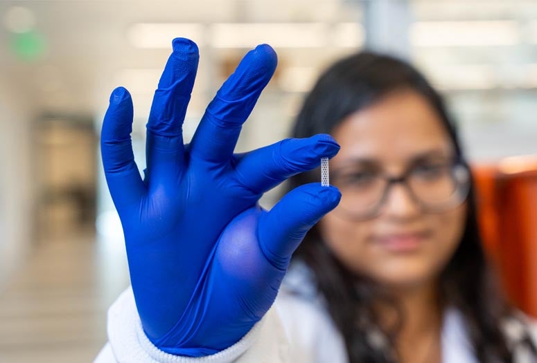 A person in blue gloves holds a tiny, patterned object, with a blurred background showing another individual in a lab setting.