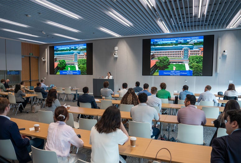 A speaker stands at a podium in a modern university lecture hall, presenting slides titled "Materials Science and NanoEngineering (MSNE)" on two large screens to an audience of university leaders and researchers seated at long wooden tables facing the front.