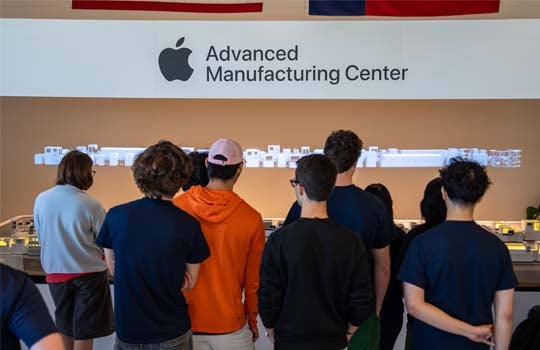 A group of students viewed from behind stands in front of a display at Apple’s Advanced Manufacturing Center, beneath a large wall sign featuring the Apple logo and center name.