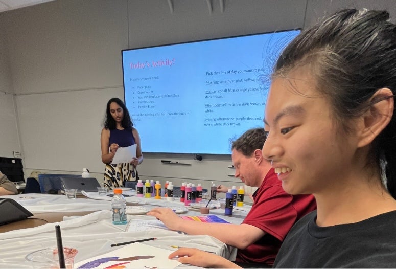 A smiling asian woman sits at a table; a woman behind her reads notes in front of a presentation