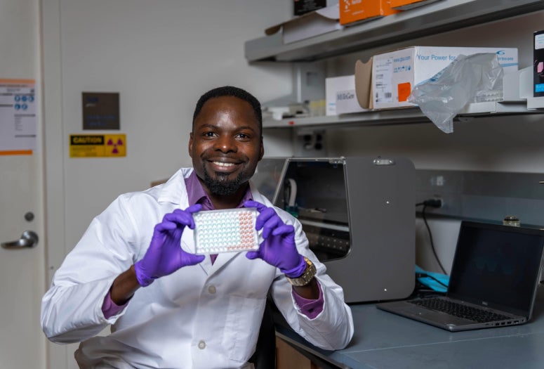 A black man in a lab coat and gloves holds up a 96 well plate while smiling
