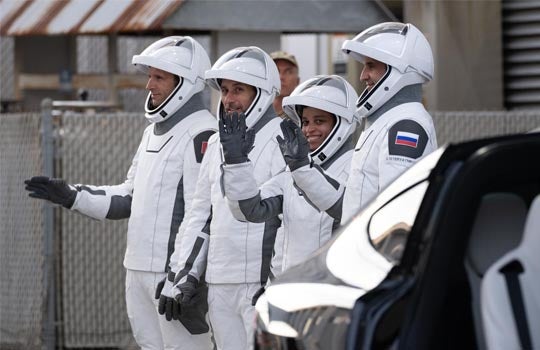 Four astronauts wearing white and black SpaceX spacesuits and white helmets walk together outdoors near a vehicle, with three waving toward the side.