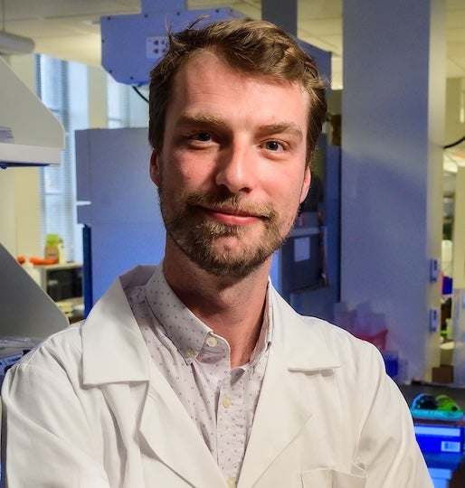 A white man stands in a lab and smiles slightly at the camera