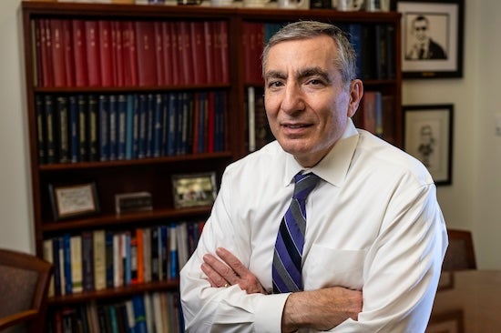 An elderly white man stands in front of a bookshelf and smiles slightly at the camera.
