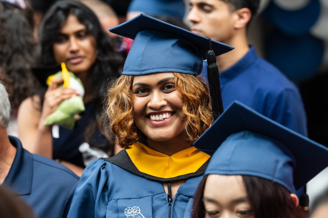 A female graduate smiles from the graduation lineup before walking across the stage to receive her diploma.