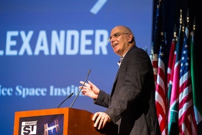A bald white man stands at a podium and gesticulates