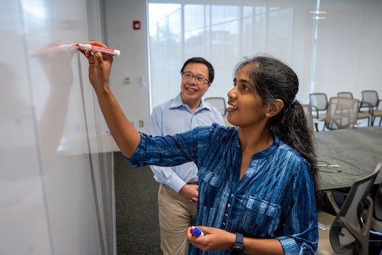 A South Asian woman draws on the white board while an East Asian man looks at what she's writing.