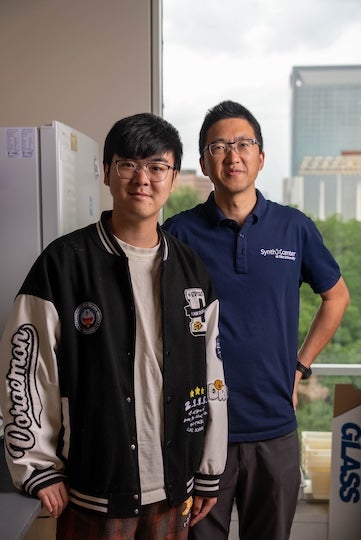 A young Asian man in a letterman jacket stands in front of a middle-aged Asian man in a polo. They are both wearing glasses.