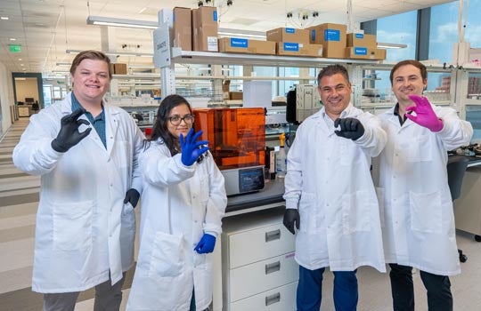 Four scientists in lab coats and gloves pose in a modern laboratory, making hand gestures while standing near equipment and shelves.