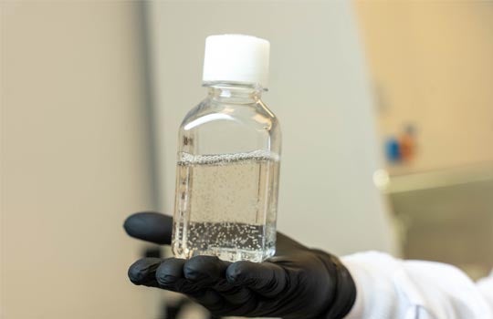 A gloved hand holds a clear bottle containing a liquid with small bubbles, likely in a laboratory setting.