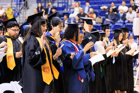 students in graduation regalia with raised hands for pledge