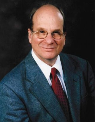 Louis Brus, an older white man in glasses and a suit, smiles at the camera
