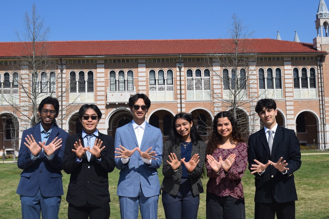 Six Rice students in making the owl sign in front of a campus building