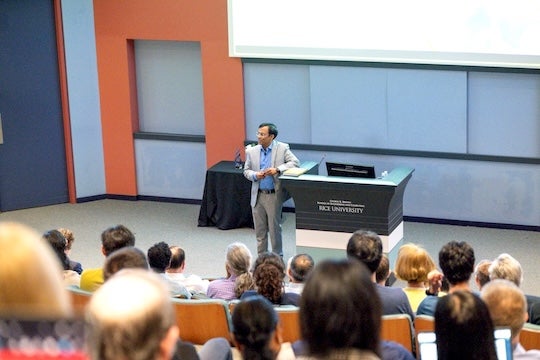 man in light gray suit beside a lectern addresses seated audience