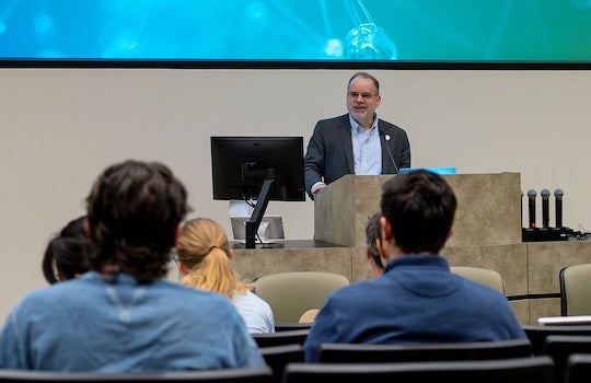 An older white man stands at a podium, talking a people seated in front of him.