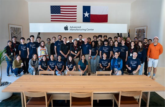A group of students stands in front of a display at Apple’s Advanced Manufacturing Center, beneath a large wall sign featuring the Apple logo and center name.