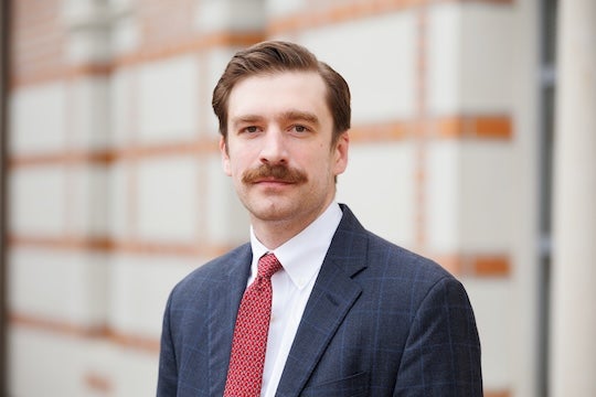 A white man with a bushy mustache stares at the camera, smiling slightly.