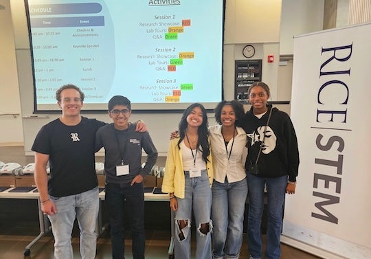 a group of five students looks at the camera, with an AV projector screen and a Rice STEM poster visible in the room behind and near them, respectively