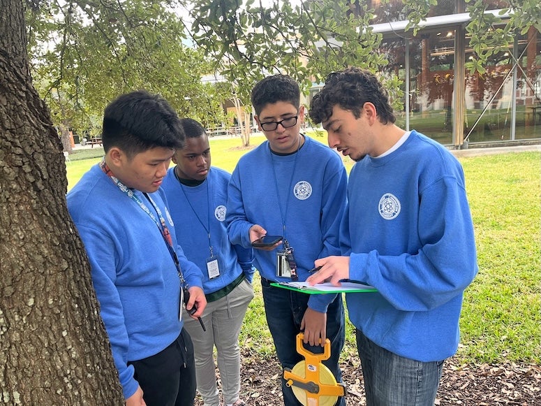 Students are standing outside, under a tree. One student has a clipboard and everyone is looking at it.