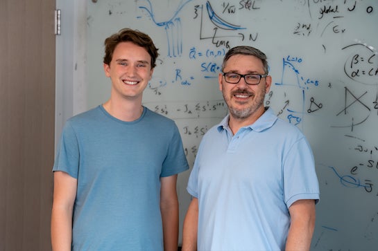 A young man and a middle aged man, both white, stand in front of a whiteboard. 