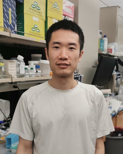 An Asian man stands in a lab and smiles slightly at the camera