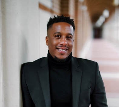 A black man leans against the wall of an outside hallway and smiles at the camera