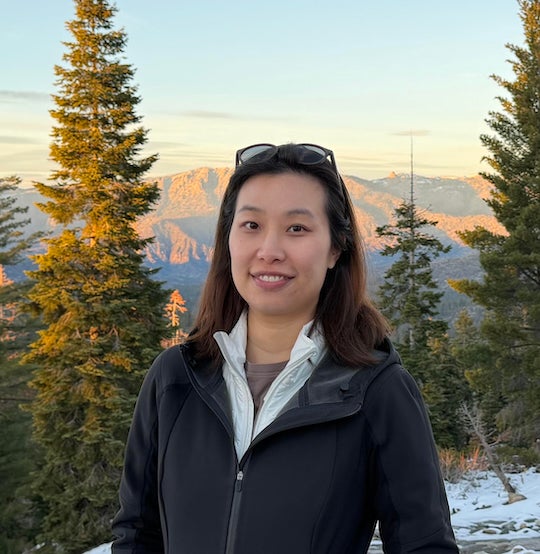 An Asian woman stands outdoors, with mountains behind her