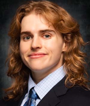 A studio headshot of a young man with long, shoulder-length wavy golden-brown hair, smiling lightly at the camera while wearing a dark suit jacket, light blue plaid dress shirt, and a blue tie.