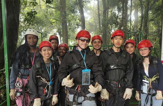 A diverse group of eight young people wearing safety helmets, harnesses, and protective gloves pose together outdoors on a platform surrounded by a lush forest.