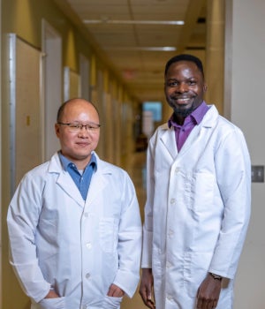 An East Asian man in a lab coat stands next to a Black man in a lab coat. They are both smiling
