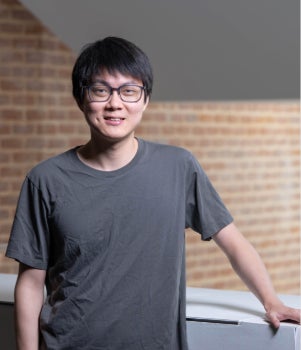 An Asian man stands in front of a glass staircase and smiles. He has a dimple. 