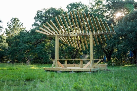 A wooden platform holds a picnic table with a leaf-shaped roof made of wooden slats