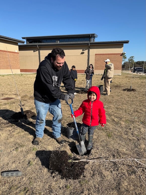 A father and son dig a hole for a tree. The four year old son is smiling at the camera.