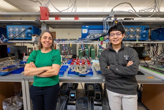 An older white woman and a young Asian man stand in front a lab bench with bottles and wires on it.